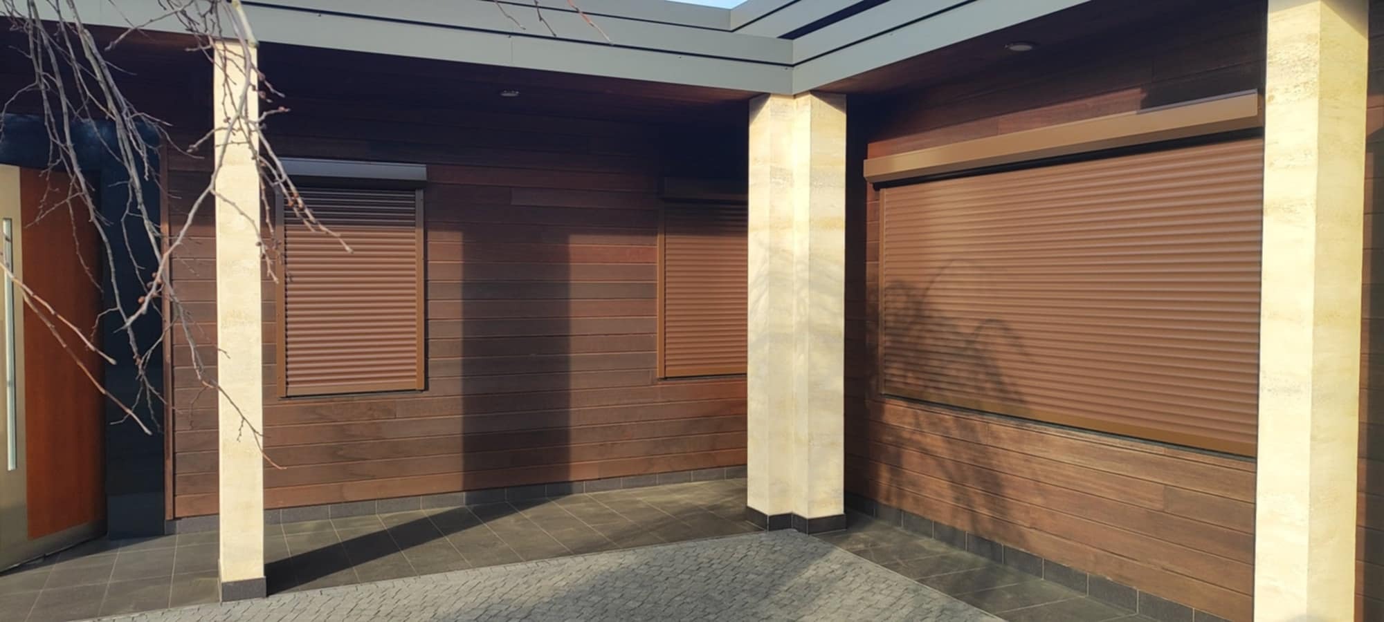 Zewnętrzne, brązowe rolety na elewacji budynku z drewnianą okładziną, widoczne kamienne filary i fragment kostki brukowej przed budynkiem.