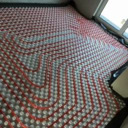 Czerwone rury ogrzewania podłogowego ułożone spiralnie na szarym podłożu z wypustkami, widoczne fragmenty czarnej folii i okno w tle.