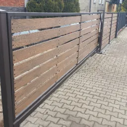 Przesuwna brama wjazdowa z poziomych desek kompozytowych w kolorze drewna, osadzona w ciemnobrązowej metalowej ramie, widok z chodnika z kostki brukowej, czerwony samochód w tle.