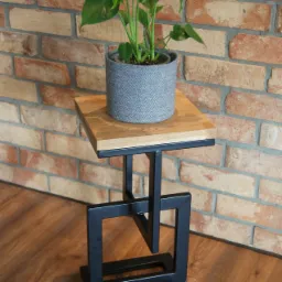 Stolik kawowy z blatem z jasnego drewna i podstawą z czarnego metalu o geometrycznym kształcie, na blacie stoi roślina w szarej doniczce, tło stanowi ściana z cegły.