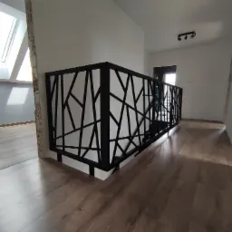 Nowoczesna balustrada metalowa z geometrycznym wzorem na antresoli, widok z góry na drewnianą podłogę, jasne wnętrze z oknem dachowym.