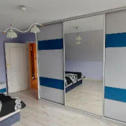 Szafa przesuwna z lustrzanymi drzwiami i niebieskimi akcentami w sypialni z fioletowymi ścianami, widoczny fragment ł&oacute;żka i drzwi.