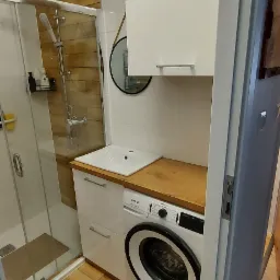 Mała łazienka z kabiną prysznicową, pralką pod blatem drewnianym i wiszącymi szafkami w kolorze białym.