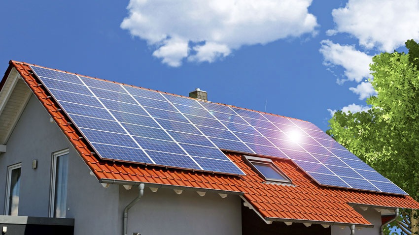 Dom jednorodzinny z szarą elewacją i czerwoną dachówką, na której zamontowane są niebieskie panele fotowoltaiczne odbijające promienie słoneczne, widoczne okno dachowe i fragment korony drzewa...