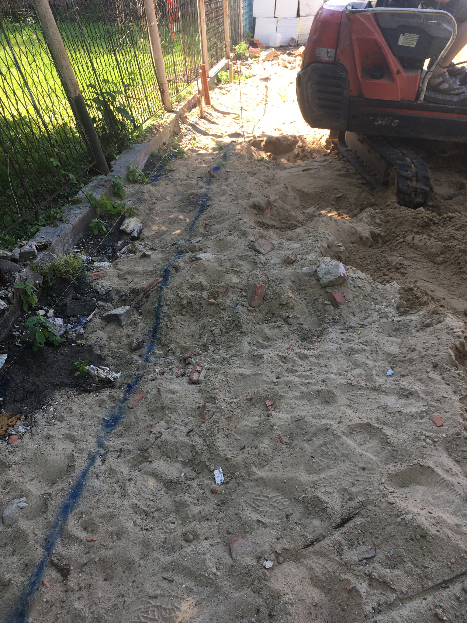 Przygotowanie terenu pod fundamenty: wykop z piaskiem, fragment koparki i wyznaczona niebieską farbą linia przyszłego fundamentu.