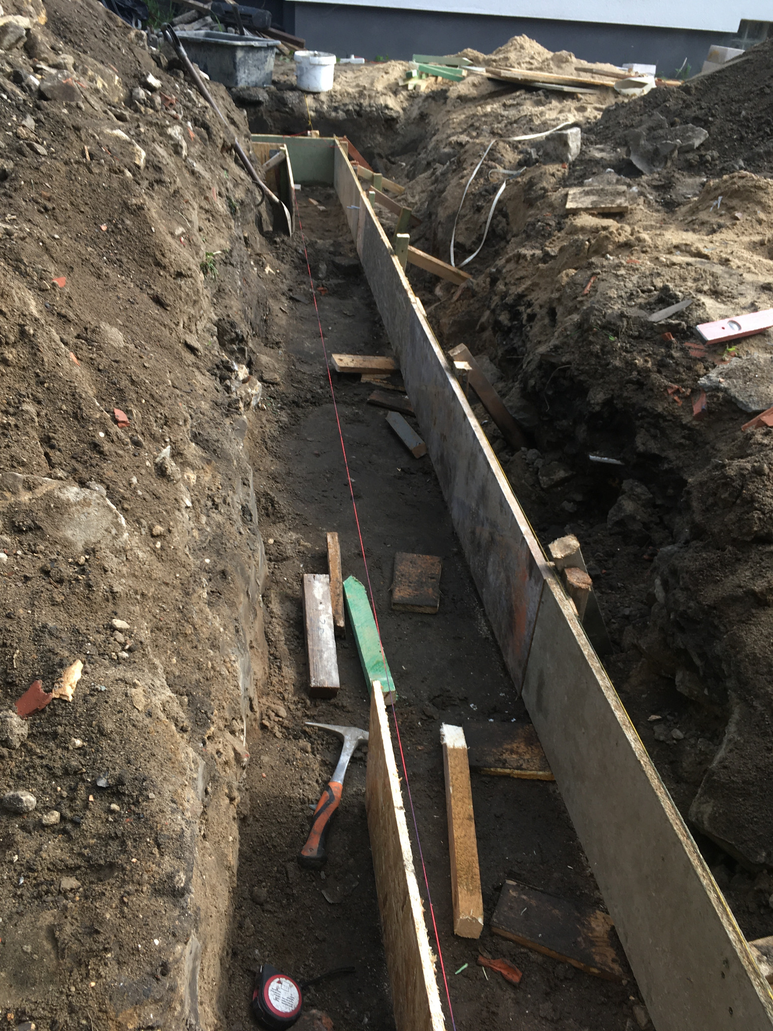 Wykop pod fundament z deskowaniem, widoczny sznurek traserski, poziomica, młotek ciesielski i miara zwijana.