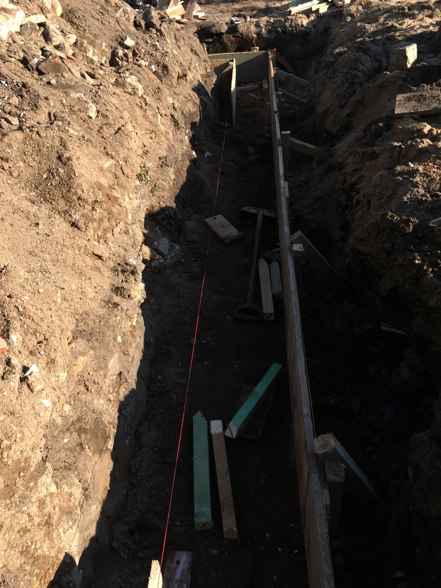 Wykop fundamentowy z drewnianym szalunkiem, widoczna poziomica sznurkowa, łopata i kliny, ziemia po bokach wykopu.
