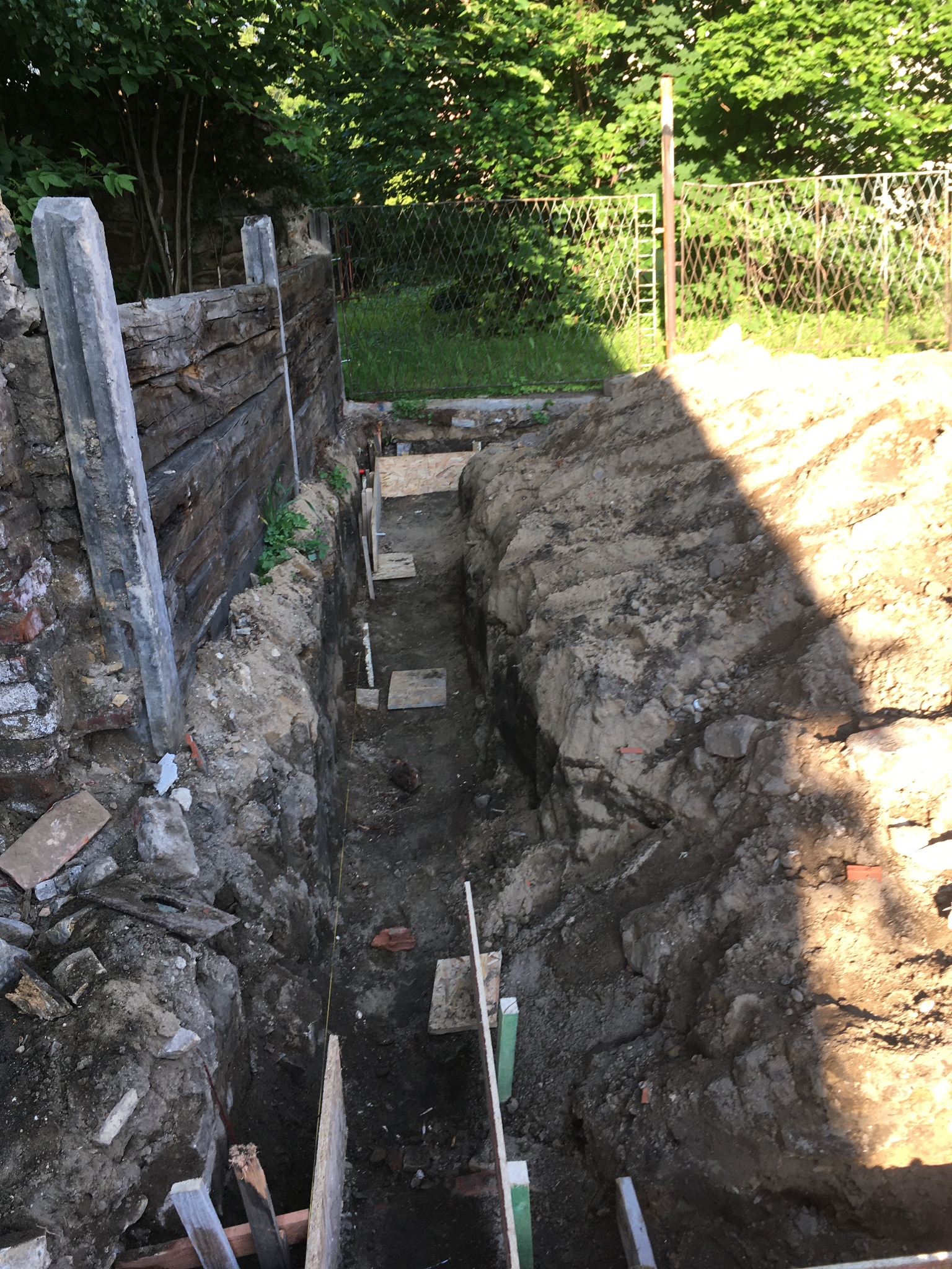 Wykop pod fundament z drewnianym szalunkiem, widoczna warstwa gruntu i fragment muru oporowego z cegły