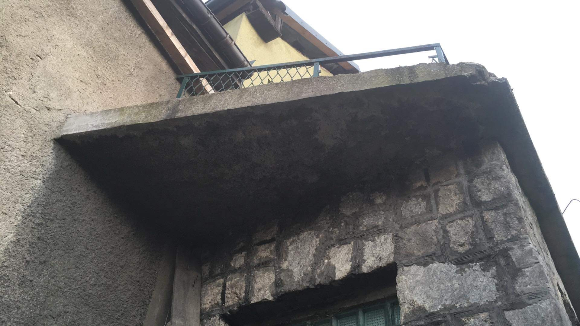 Niedokończony betonowy balkon z metalową balustradą, widoczna surowa ściana z kamieni i fragment okna z zielonymi szybami, elewacja budynku w stanie surowym.