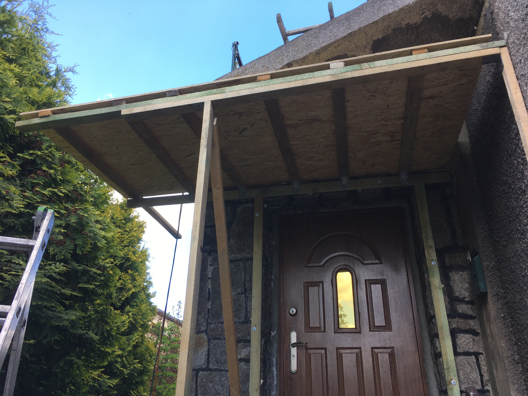 Drewniana konstrukcja z płyt OSB budowana nad wejściem do domu, widoczna drabina oparta o ścianę, fragment iglastego drzewa w tle.