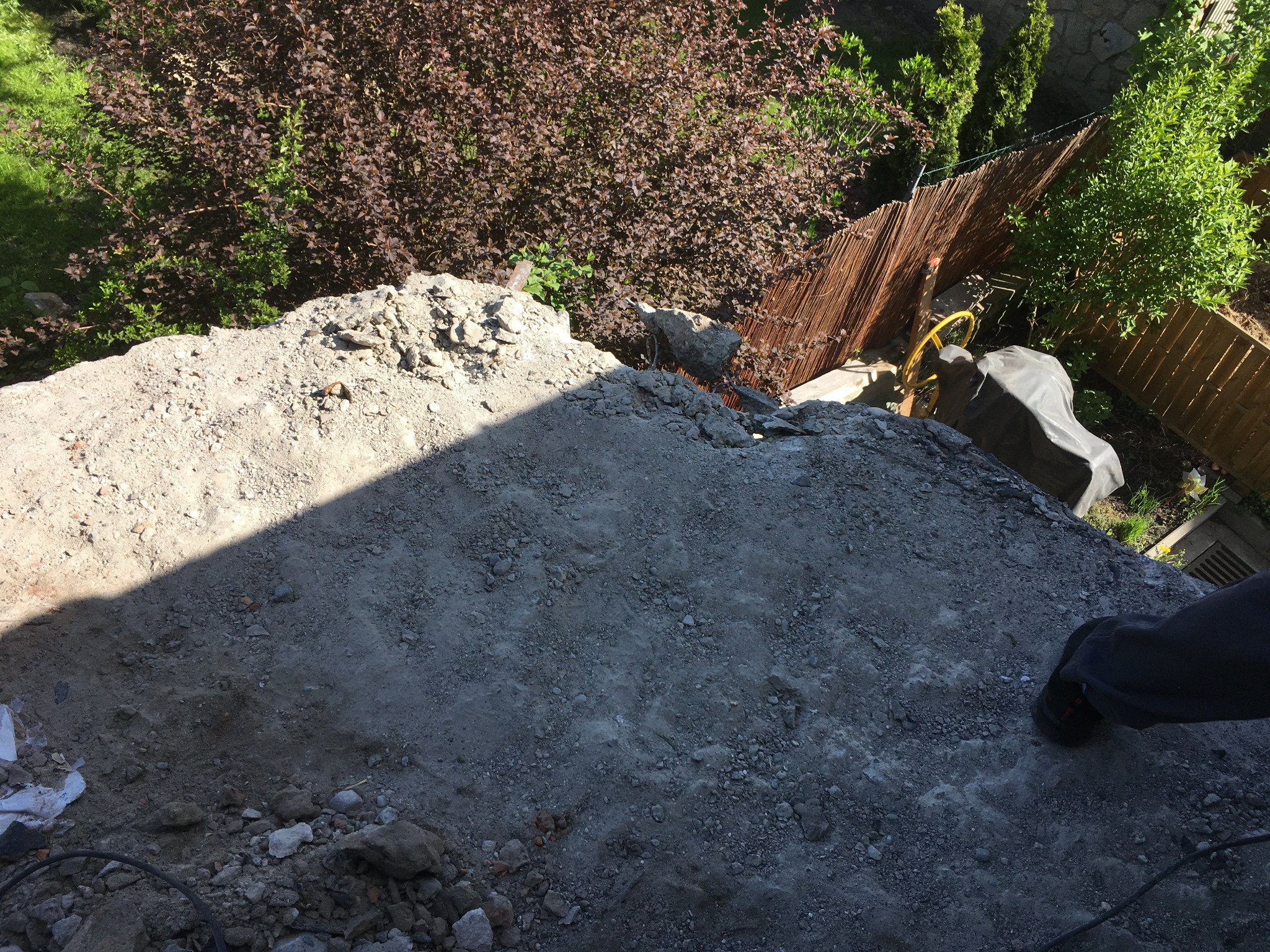 Niedokończona betonowa konstrukcja w ogrodzie, widok z góry, fragment stopy pracownika w prawym dolnym rogu, w tle roślinność i ogrodzenie.