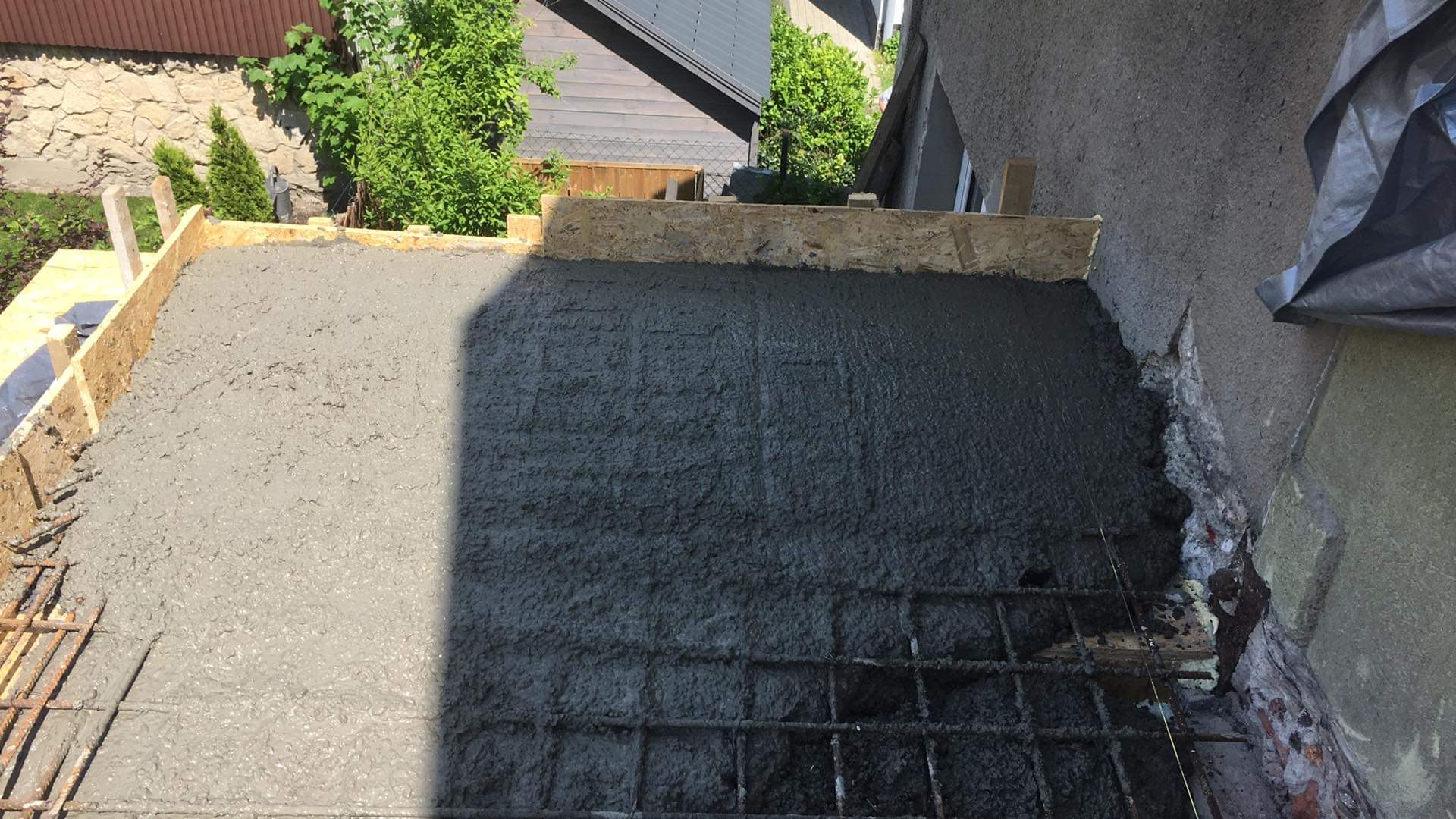 Świeżo wylany beton na zbrojeniu, otoczony drewnianym szalunkiem, przygotowany do dalszych prac budowlanych na zewnątrz budynku.