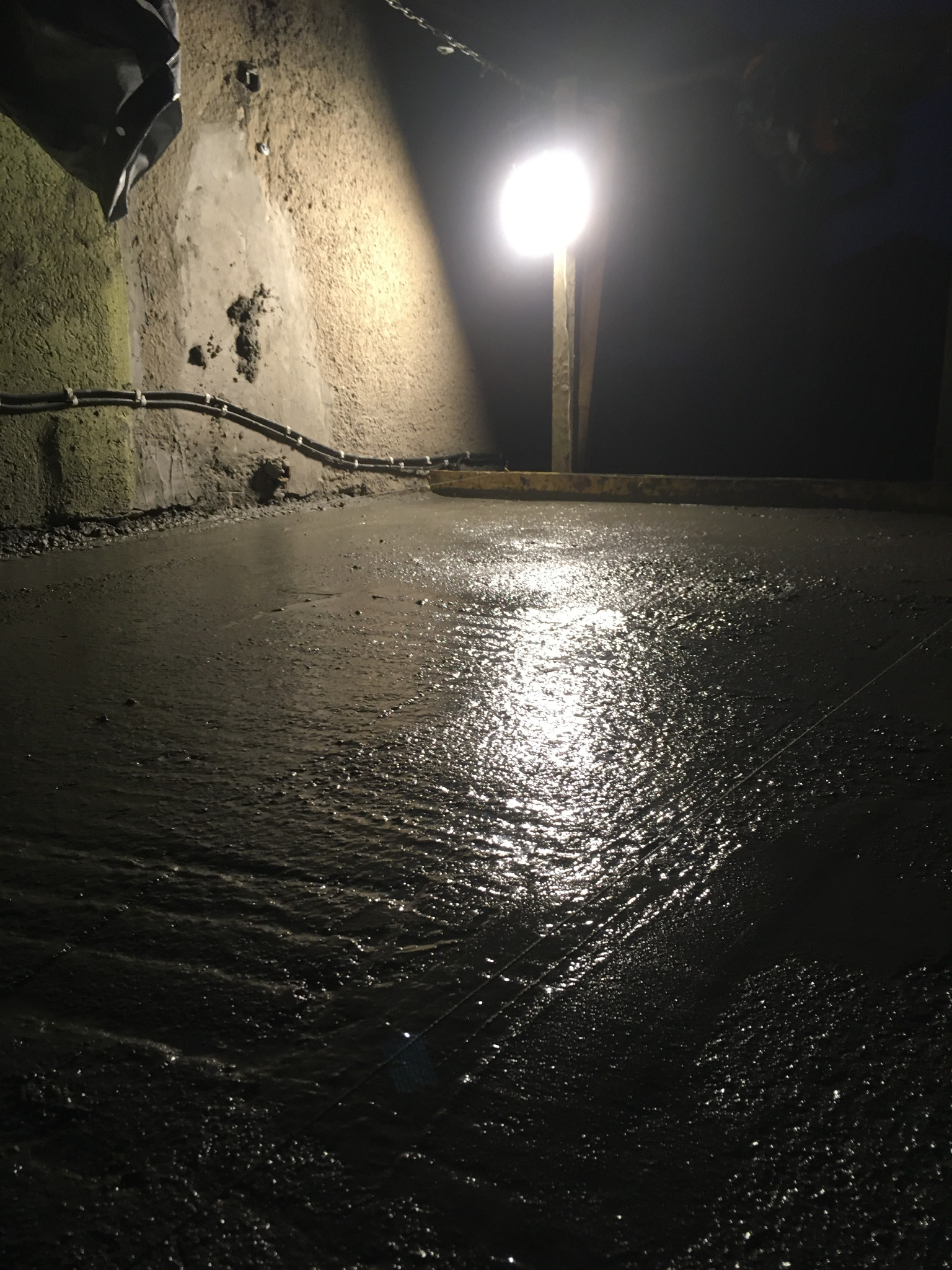 Świeżo wylany, mokry beton na podłodze w słabo oświetlonym pomieszczeniu z widocznymi przewodami elektrycznymi przy ścianie i sznurkiem wyznaczającym poziom.