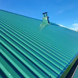 Świeżo pomalowany, zielony dach z blachy trapezowej, z widocznym kominem i błękitnym niebem w tle.