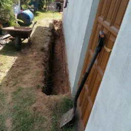 Wykopany wąski rów w ziemi wzdłuż ściany budynku, obok oparta łopata, w tle taczka i niebieska beczka.