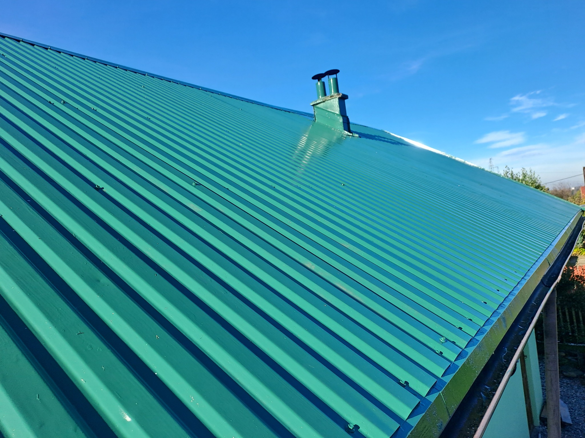 Świeżo pomalowany, zielony dach z blachy trapezowej, z widocznym kominem i błękitnym niebem w tle.