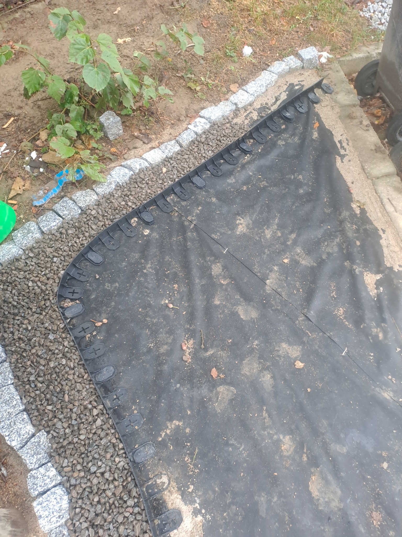 Czarna agrowłóknina rozłożona na ziemi, obok kamienny obrzeże i małe kamyczki, widoczne elementy roślinności