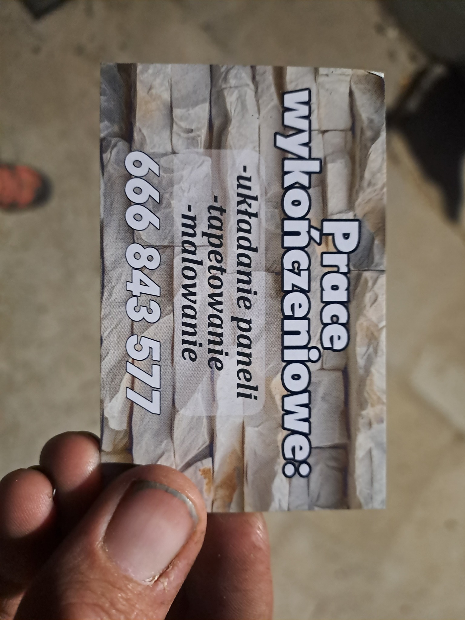 Wizytówka firmy remontowej z numerem telefonu i zakresem usług: układanie paneli, tapetowanie, malowanie, na tle imitacji kamiennej ściany.