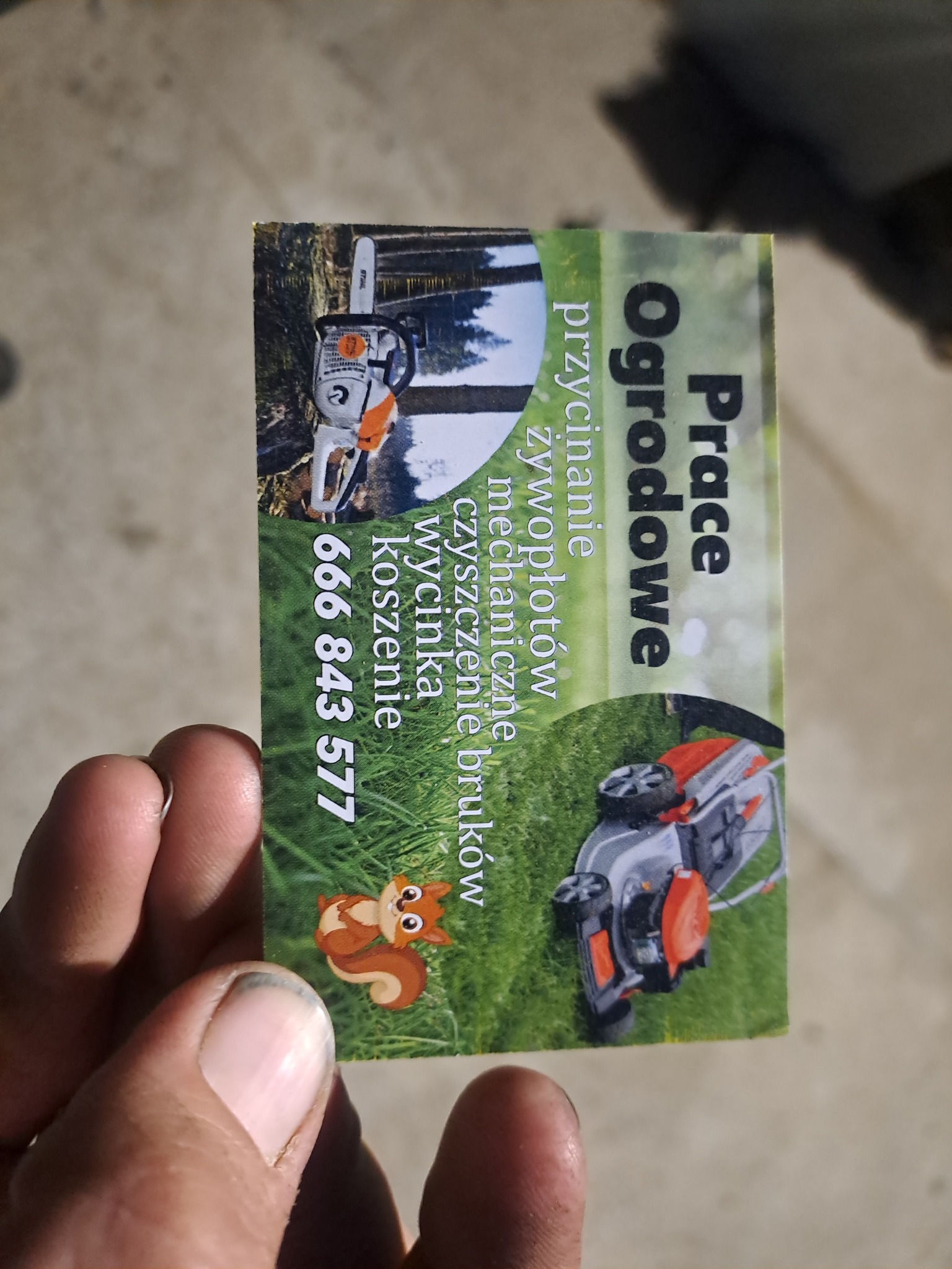 Przybliżenie wizytówki z napisem 'Prace Ogrodowe', ilustracją piły łańcuchowej, kosiarki do trawy i wiewiórki, trzymanej w palcach