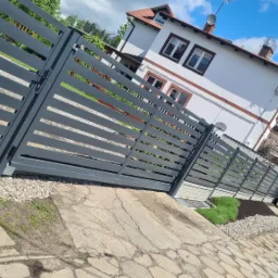 Nowoczesne, szare ogrodzenie panelowe z poziomymi szczeblami przed domem z białymi ścianami i brązowymi obramowaniami okien, widoczne kostki brukowe i fragment trawnika.