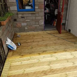 Nowo wybudowany drewniany taras z widocznymi workami z materiałami budowlanymi i narzędziami, przylegający do ściany z kamienia i otwartych drzwi do garażu z motocyklem w środku.