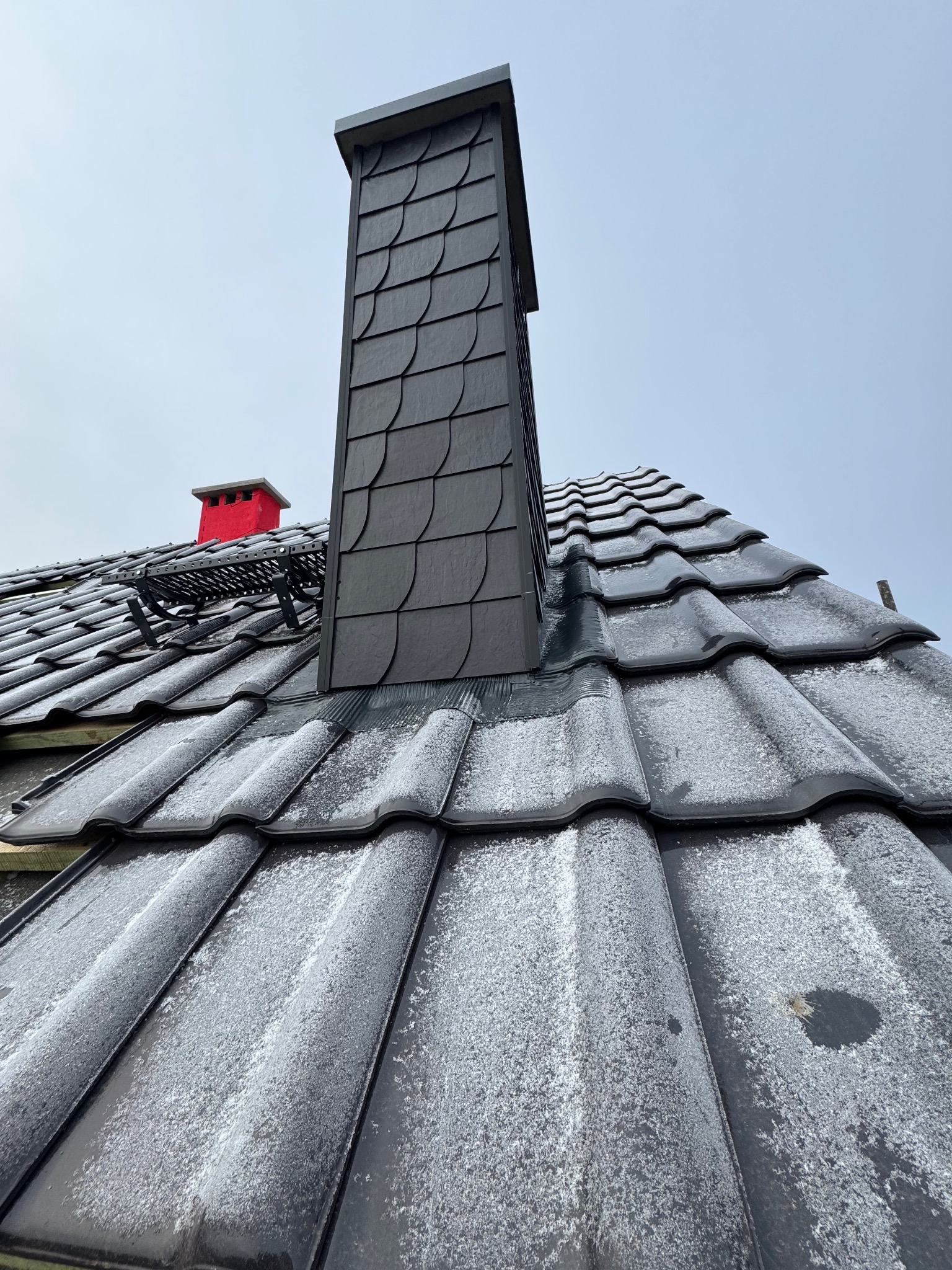 Czarny komin z łupka na dachu pokrytym błyszczącą dachówką karpiówką, lekko oszronioną. W tle czerwony komin i drabinka kominiarska. Ujęcie z dołu.