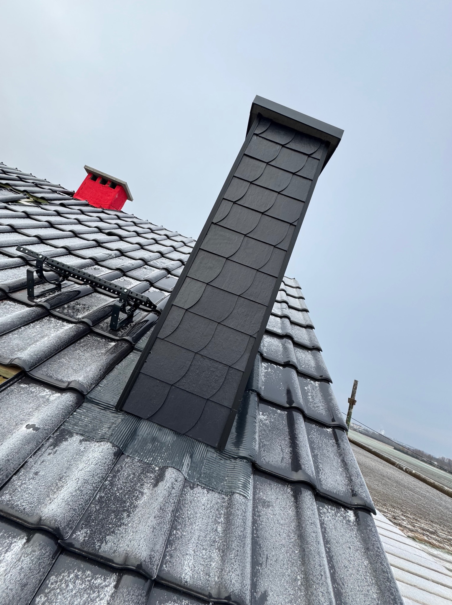 Czarny komin z łupka na dachu pokrytym szarą dachówką ceramiczną, częściowo oszronioną. Widoczny czerwony kominek wentylacyjny i drabinka dachowa.