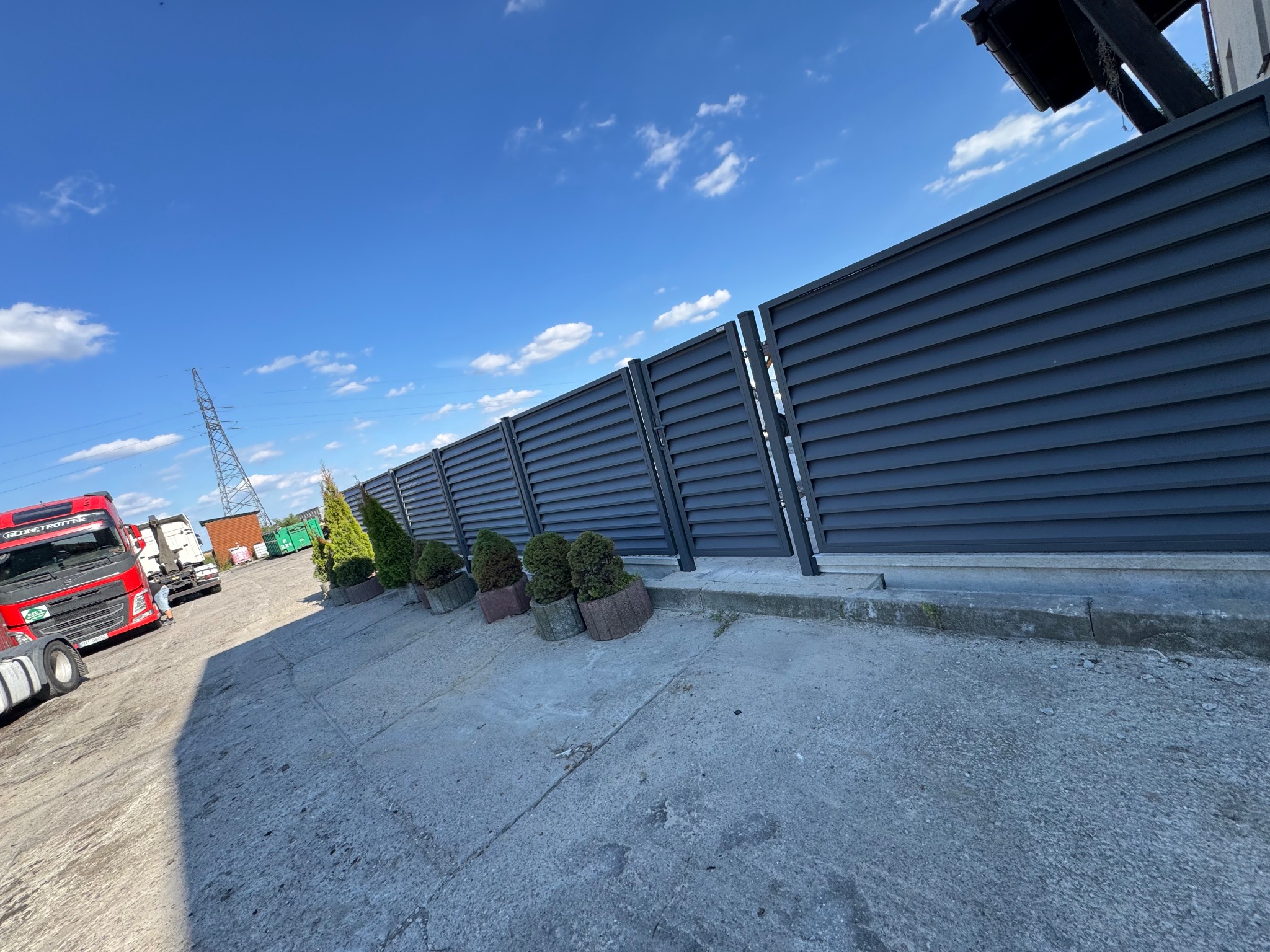 Nowoczesne, poziome ogrodzenie panelowe w kolorze antracytowym, z nasadzeniami z tuj i kulistych krzewów. Widok z perspektywy żabiej, błękitne niebo w tle.