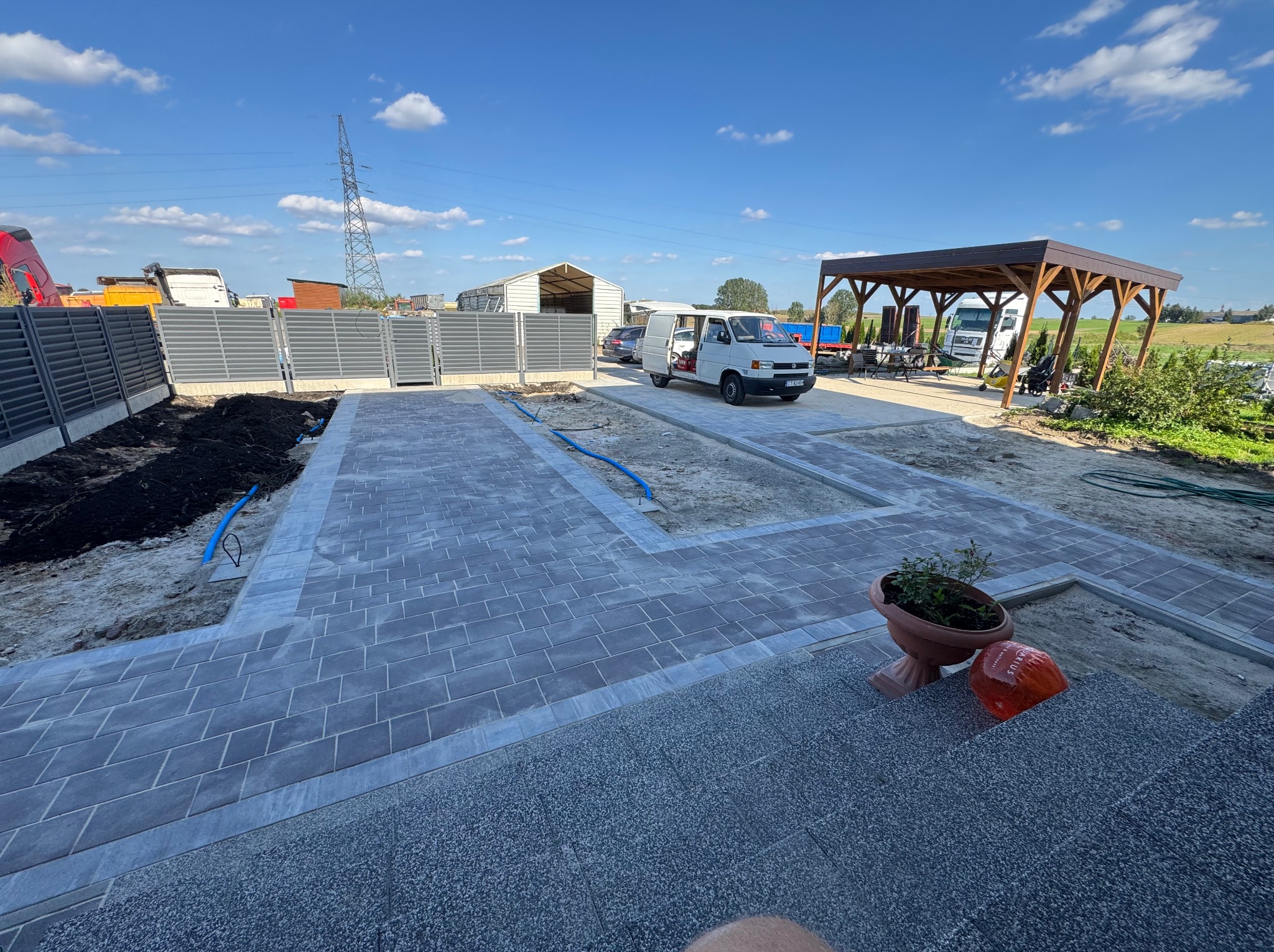 Nowa, szara kostka brukowa ułożona na podjeździe, z widocznymi fragmentami ziemi i niebieskimi rurami. W tle biały bus i szare ogrodzenie panelowe.