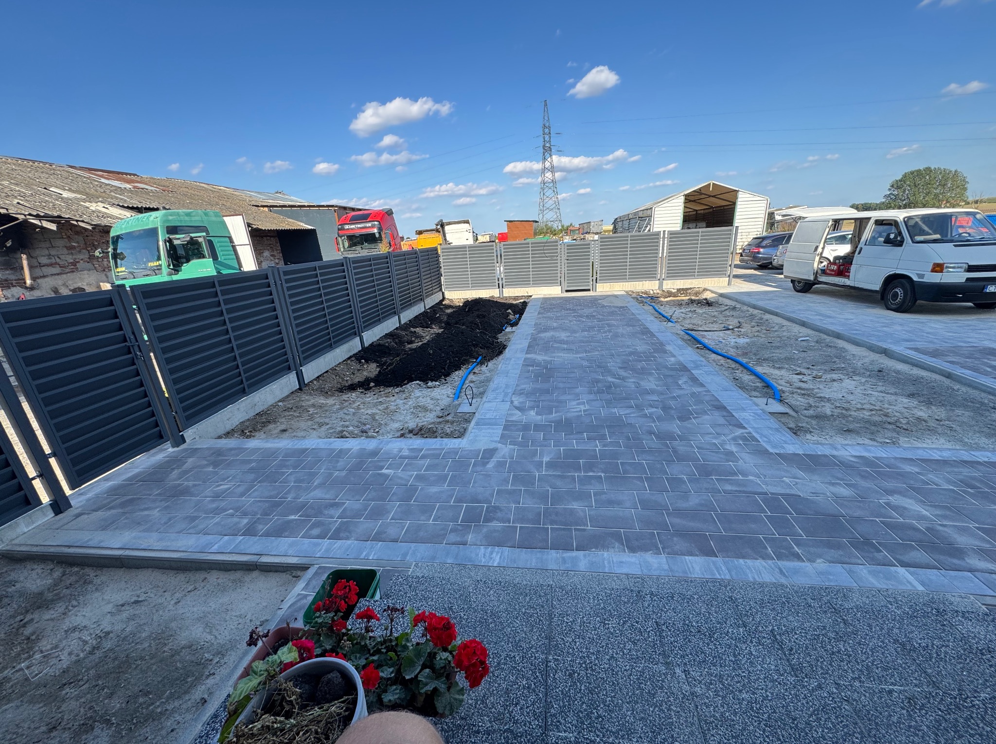 Nowoczesny chodnik z szarej kostki brukowej i metalowe ogrodzenie panelowe na posesji w Sztumie. Widoczne przygotowane rabaty i pojazdy w tle.