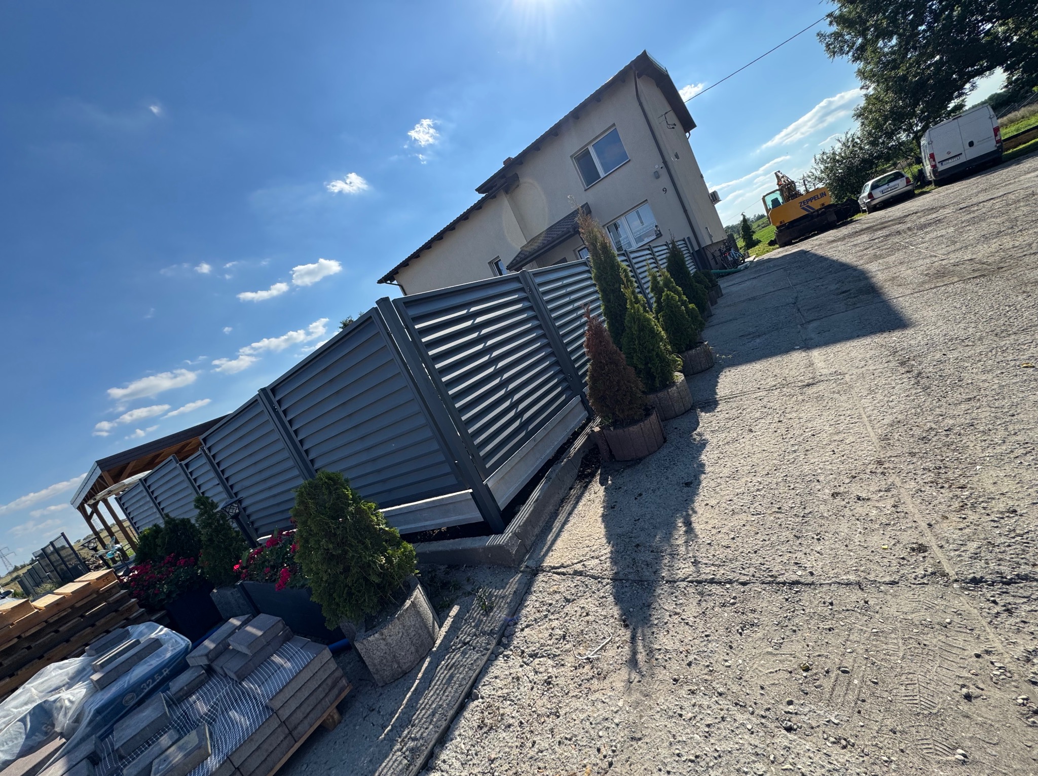 Nowoczesne ogrodzenie panelowe w kolorze grafitowym, z nasadzeniami z tuj i kwiatów w donicach. W tle dom jednorodzinny i błękitne niebo z chmurami.