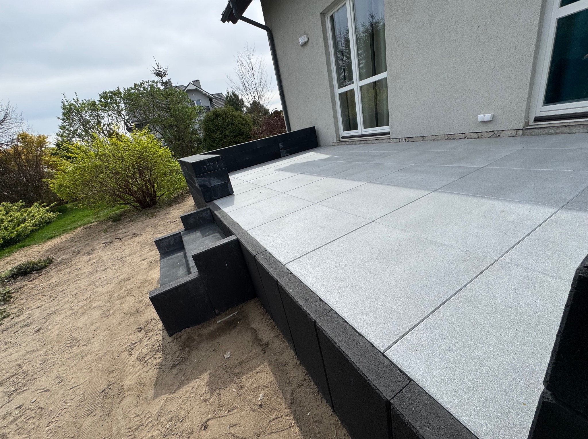 Nowoczesny taras z szarych płyt, czarne obrzeża i schody, jasna elewacja budynku w tle, widok z dołu, świeżo ułożona nawierzchnia.