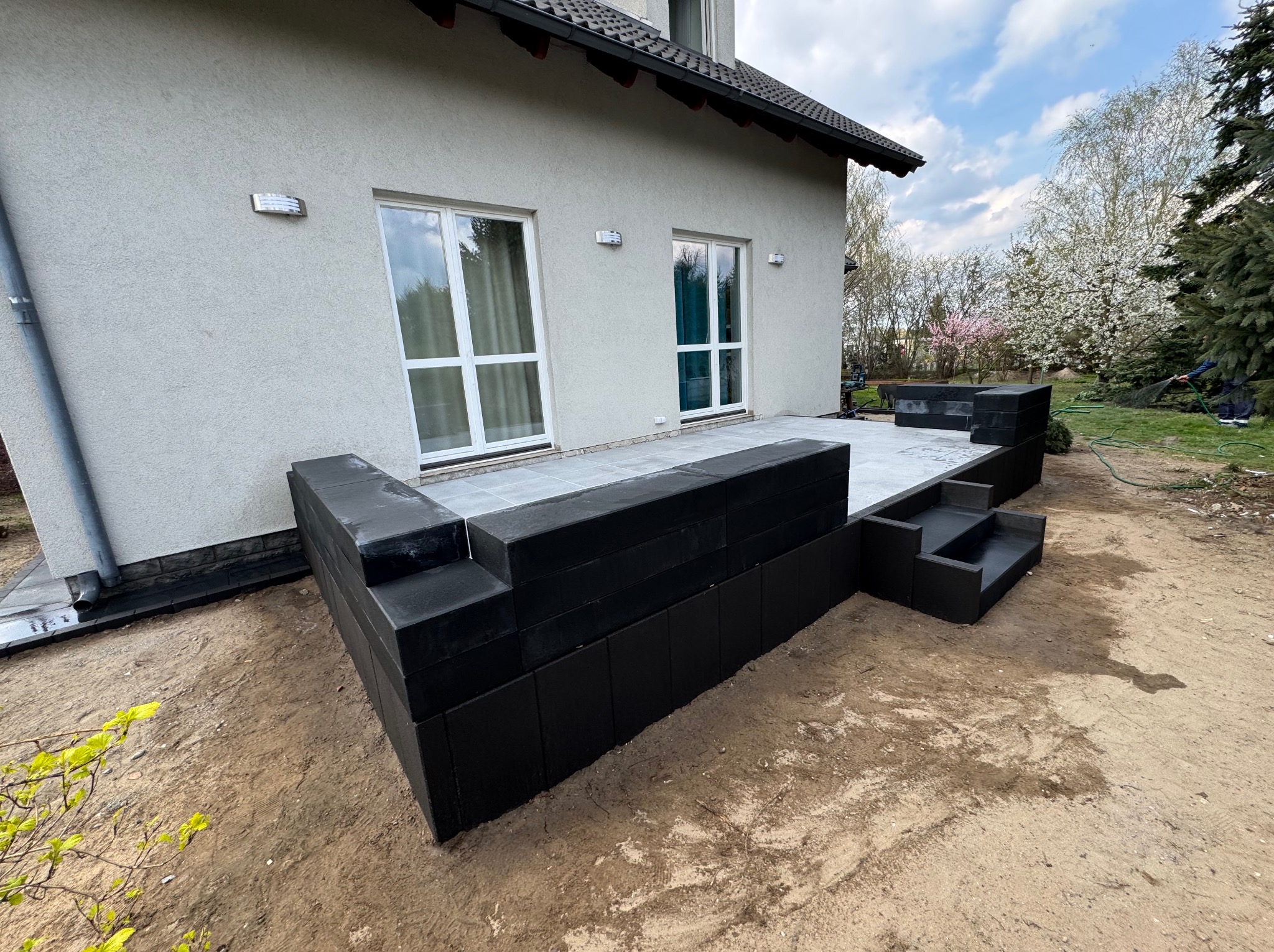 Nowoczesny taras z czarnych, modułowych elementów i szarych płyt, przylegający do jasnego domu z dwoma oknami. W tle kwitnące drzewa i trawnik.