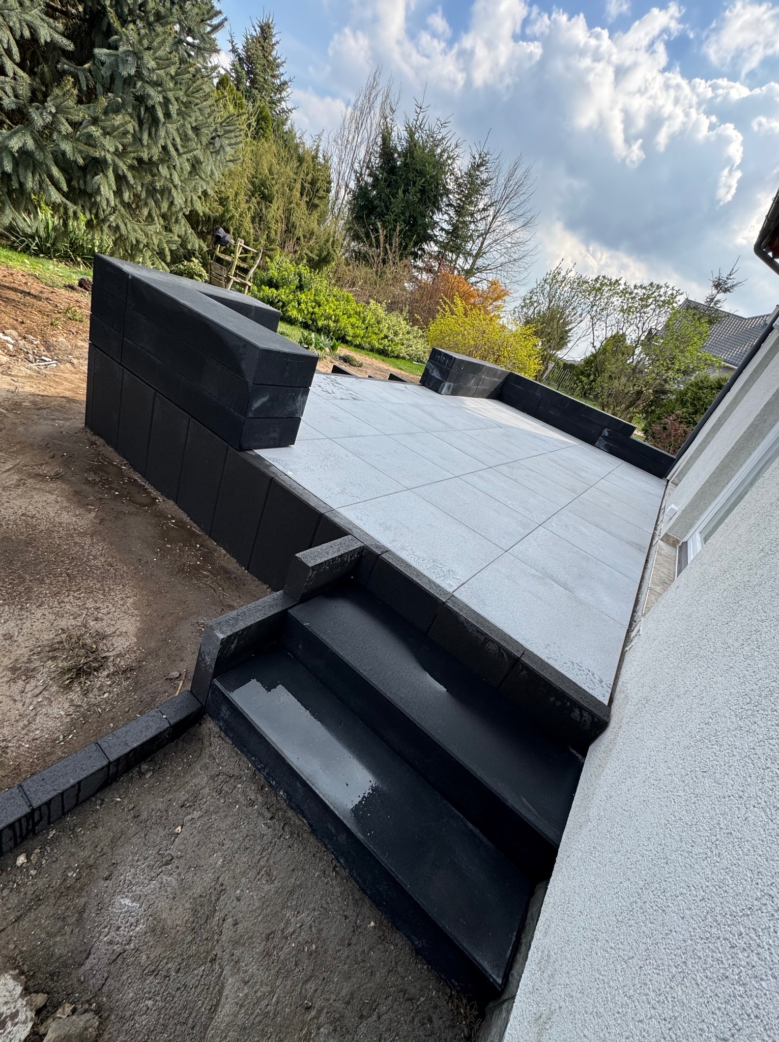 Nowoczesny taras z jasnoszarej kostki, obrzeża z czarnych elementów, schody wejściowe. Widok na ogród i błękitne niebo z chmurami. Minimalistyczny design.