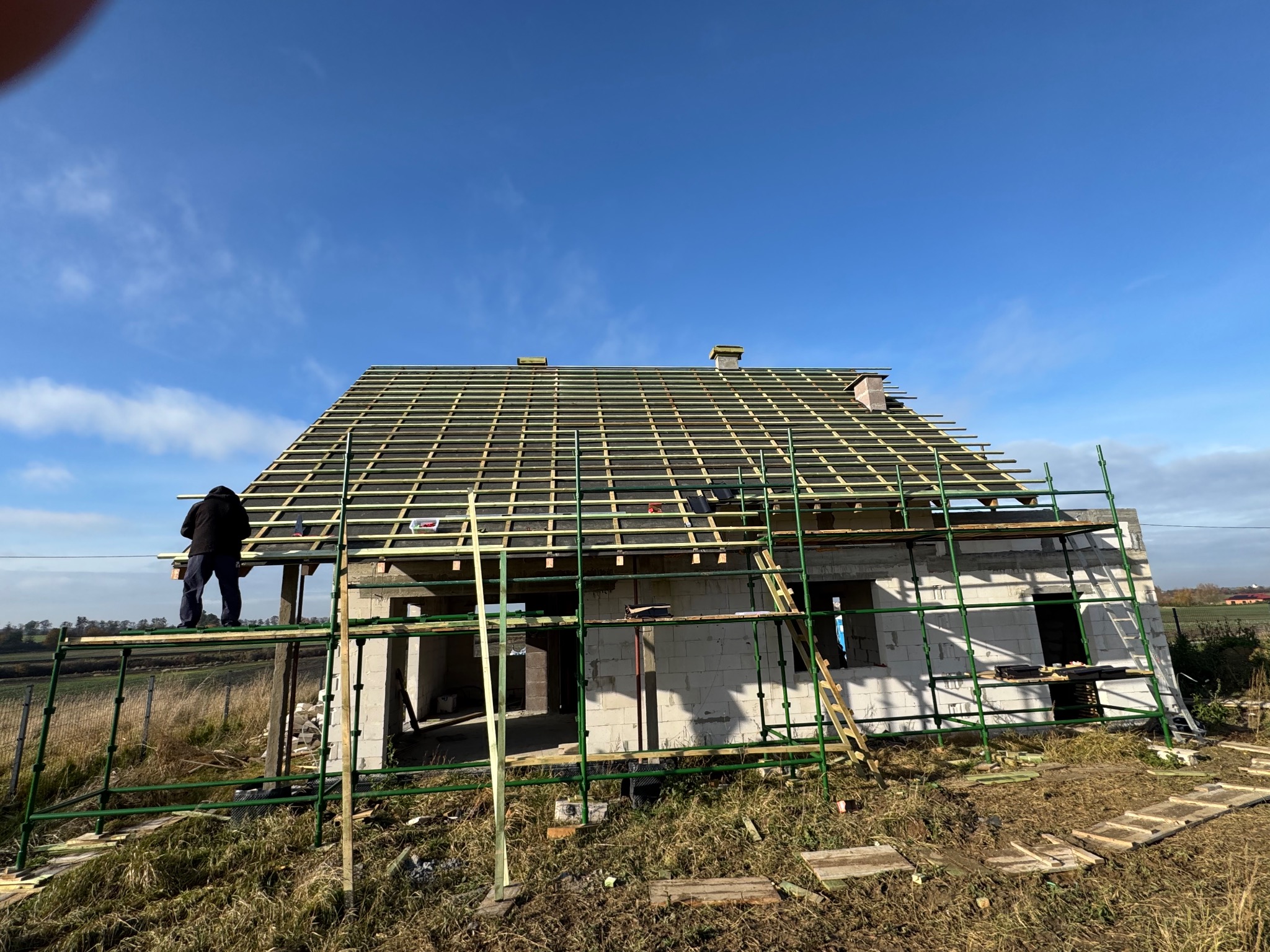 Budowa domu w toku: szkielet dachu z łatami, białe ściany z pustaków, rusztowania i pracownik na wysokości. Błękitne niebo w tle.