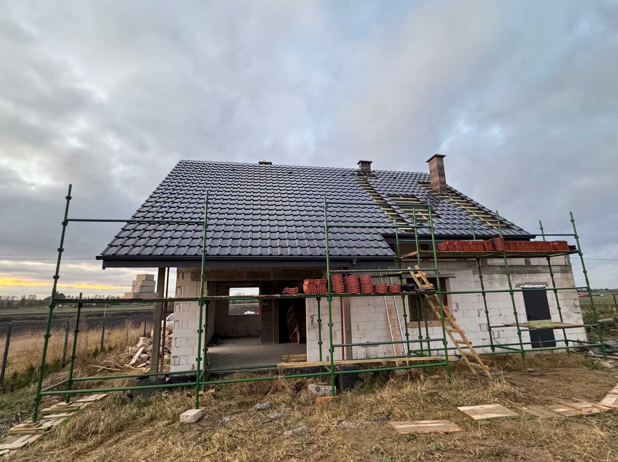 Budowa domu jednorodzinnego w trakcie realizacji: częściowo pokryty dach czarną dachówką, rusztowania, cegły, widok na okolicę w pochmurny dzień.