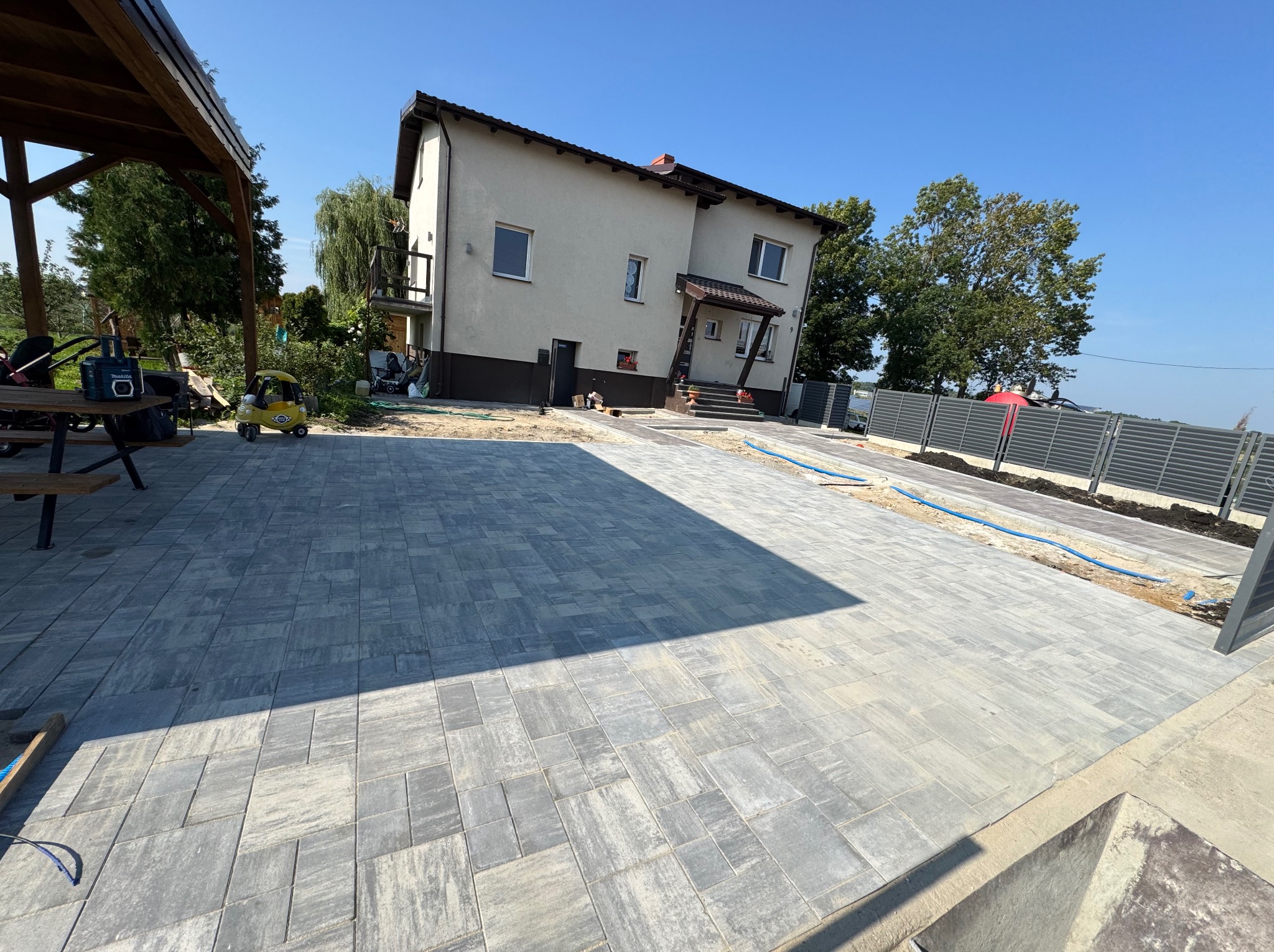 Ułożona kostka brukowa na podjeździe w Czerninie, widok na dom i ogrodzenie. Szare, prostokątne płyty tworzą wzór. Częściowo zacieniony obszar.