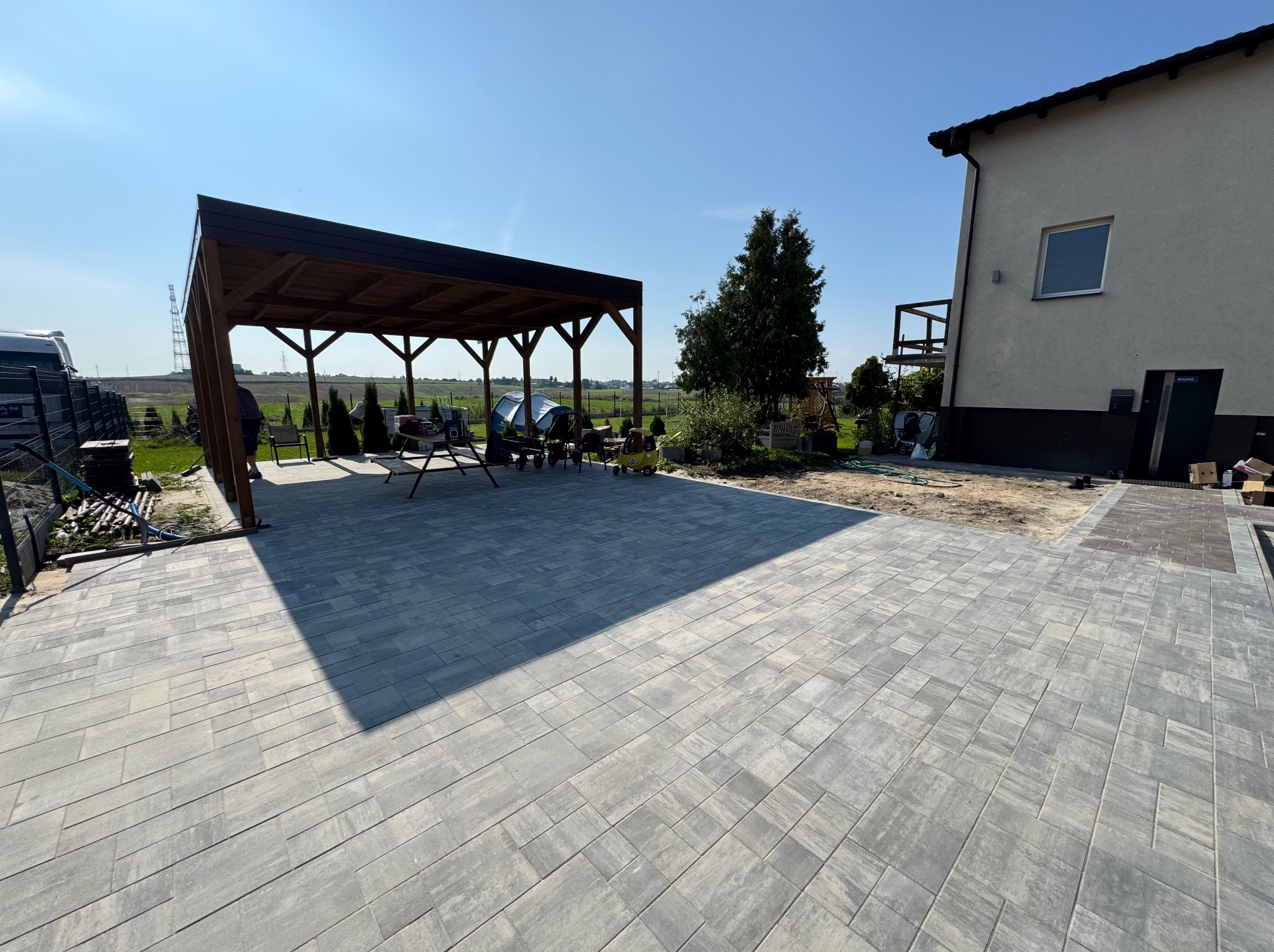 Ułożona kostka brukowa w szarym odcieniu, zadaszenie drewniane, dom z oknem i balkonem w tle, słoneczny dzień. Widoczne maszyny budowlane i elementy ogrodu.