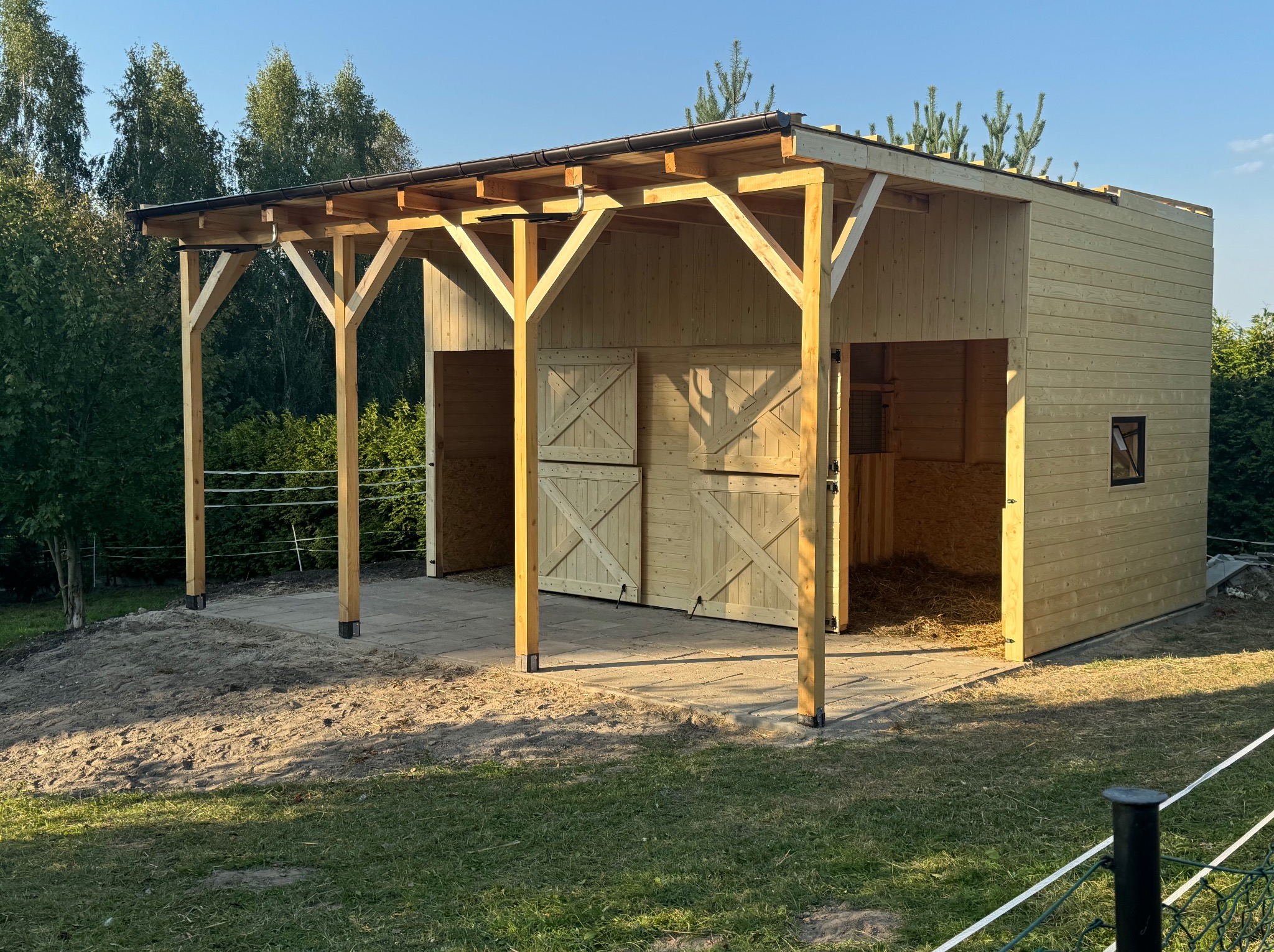 Drewniana konstrukcja z zadaszeniem i dwuskrzydłowymi drzwiami, prawdopodobnie stajnia lub wiata, na tle zieleni i błękitnego nieba w słoneczny dzień.