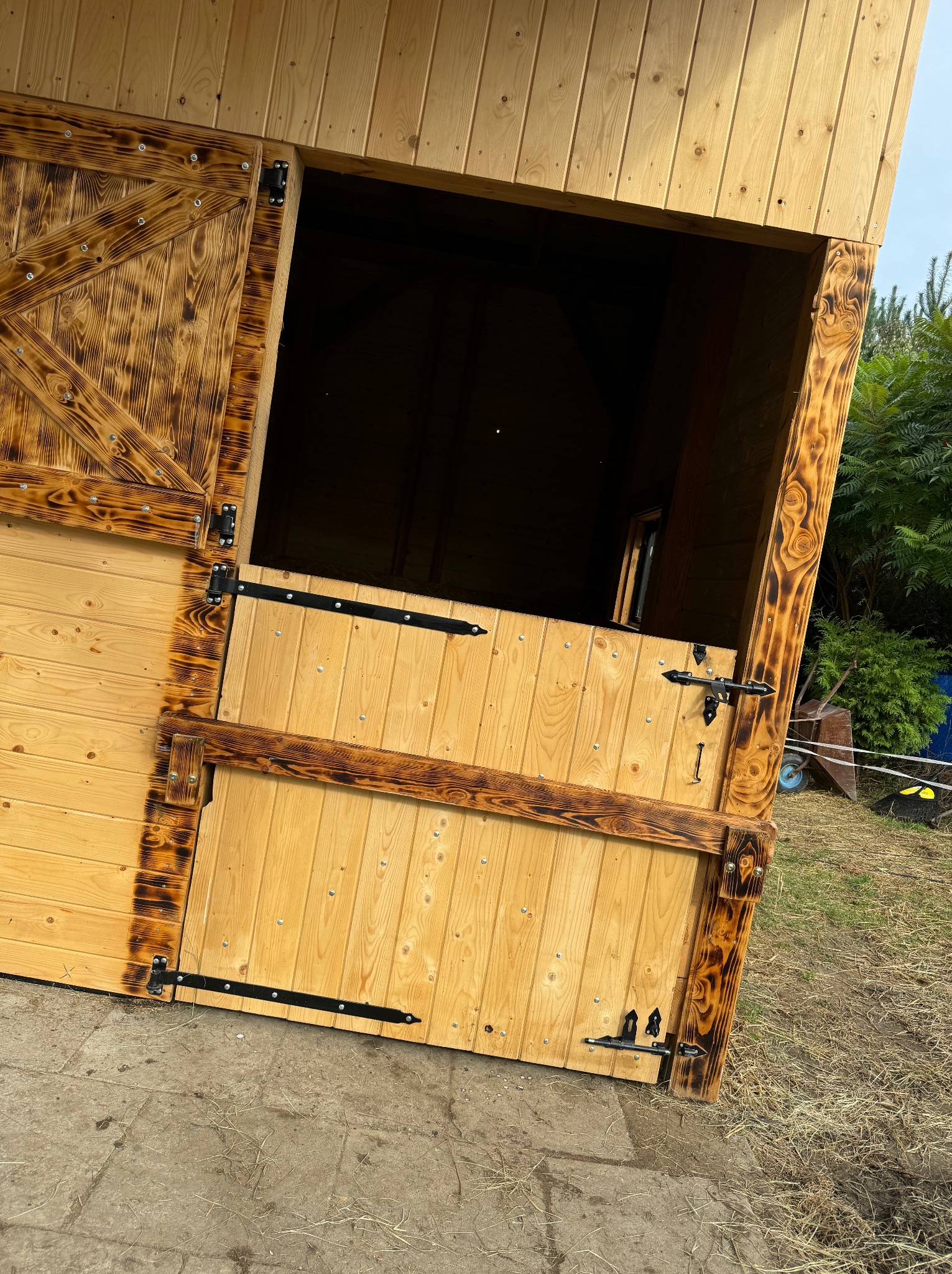 Drewniana konstrukcja z otwieranymi drzwiami, wykończona techniką opalania drewna, z widocznymi zawiasami i zasuwką, na zewnątrz, z zielenią w tle.