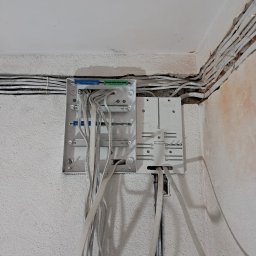 Roman Ławniczak - Rozdzielnica elektryczna z podłączonymi przewodami na nierównej, białej ścianie. Widoczne kable ułożone wzdłuż ściany. Ujęcie z bliska, ukazujące detale instalacji.