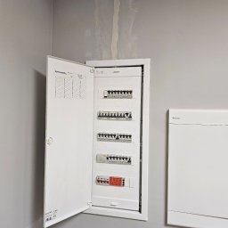 Roman Ławniczak - Otwarta, biała skrzynka rozdzielcza z bezpiecznikami na szarej ścianie. Widoczne elementy instalacji i schemat. Minimalistyczny design, neutralne oświetlenie.