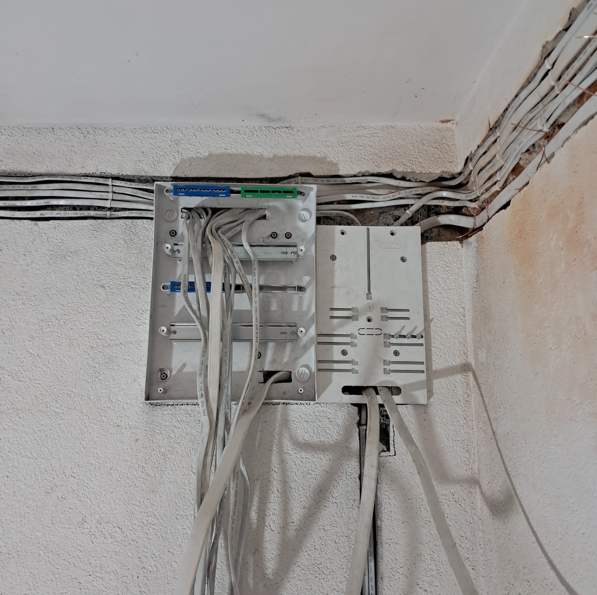 Rozdzielnica elektryczna z podłączonymi przewodami na nierównej, białej ścianie. Widoczne kable ułożone wzdłuż ściany. Ujęcie z bliska, ukazujące detale instalacji.