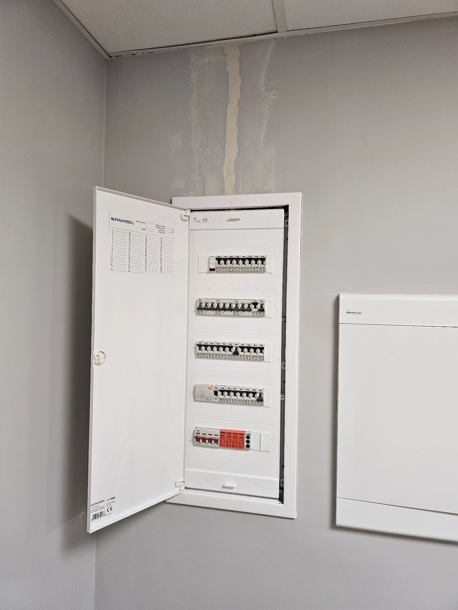 Otwarta, biała skrzynka rozdzielcza z bezpiecznikami na szarej ścianie. Widoczne elementy instalacji i schemat. Minimalistyczny design, neutralne oświetlenie.