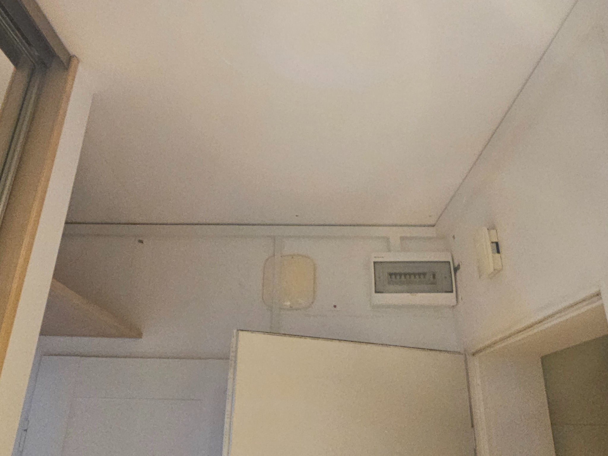 Biała skrzynka rozdzielcza z bezpiecznikami zamontowana na ścianie wewnątrz pomieszczenia. Widoczne przewody i elementy instalacji.