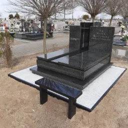 Czarny, polerowany pomnik z granitu z ławką na cmentarzu w Toruniu, otoczony żwirem i innymi nagrobkami.