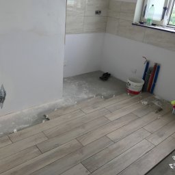 REMONTER Firma transportowo-budowlana Krzysztof Gleba - Układanie podłogi z płytek imitujących drewno w łazience, widoczne krzyżyki dystansowe, wiadro z zaprawą i narzędzia glazurnicze.