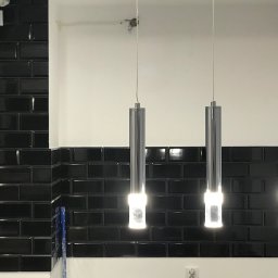 REMONTER Firma transportowo-budowlana Krzysztof Gleba - Błyszczące, czarne płytki ułożone w cegiełkę na ścianie, oświetlone przez trzy nowoczesne, chromowane lampy wiszące z białym światłem.