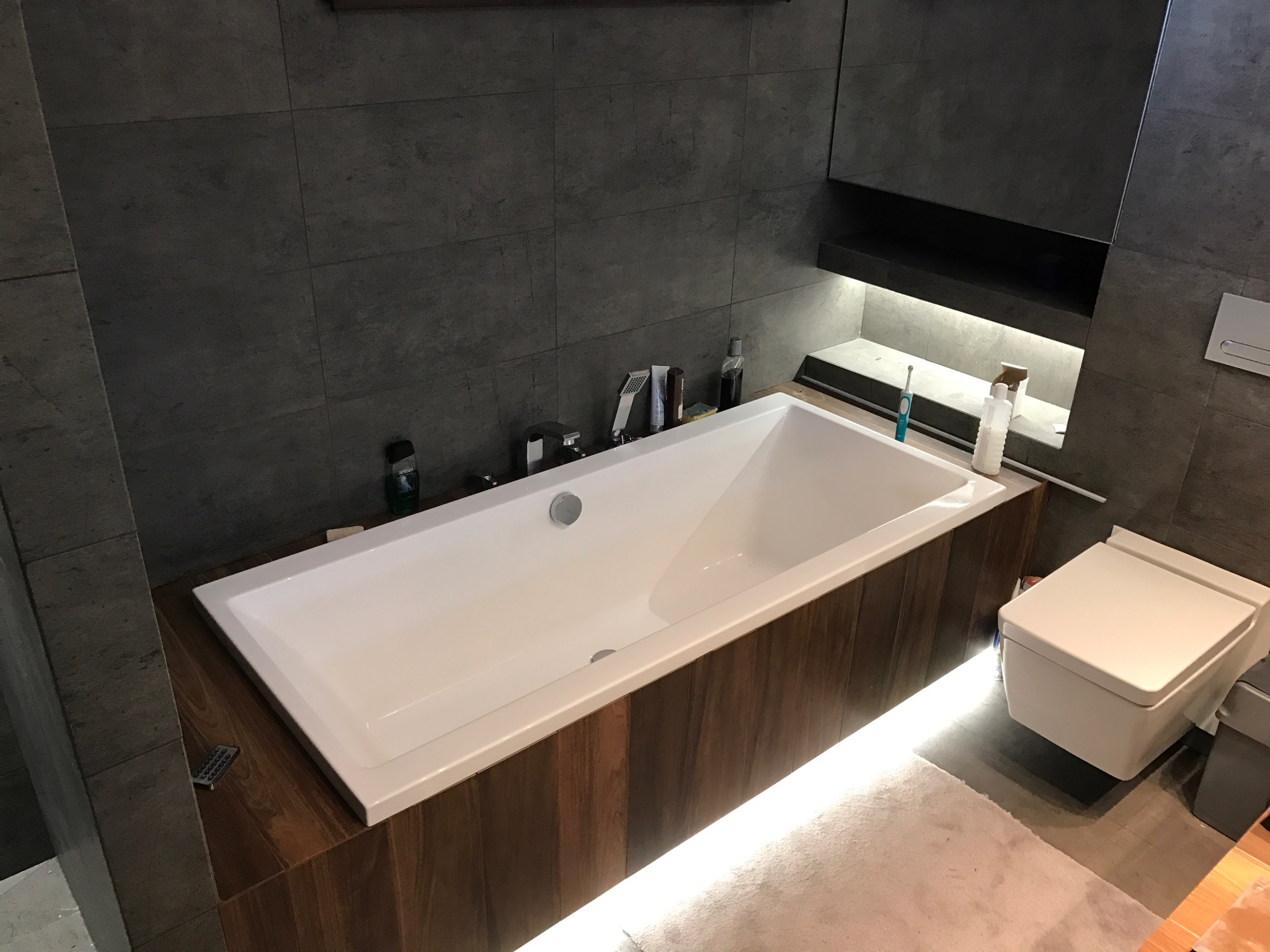 Nowoczesna łazienka z wanną obudowaną drewnopodobnymi panelami, podświetlona listwą LED, szare płytki na ścianach i podłodze, minimalistyczna toaleta podwieszana.