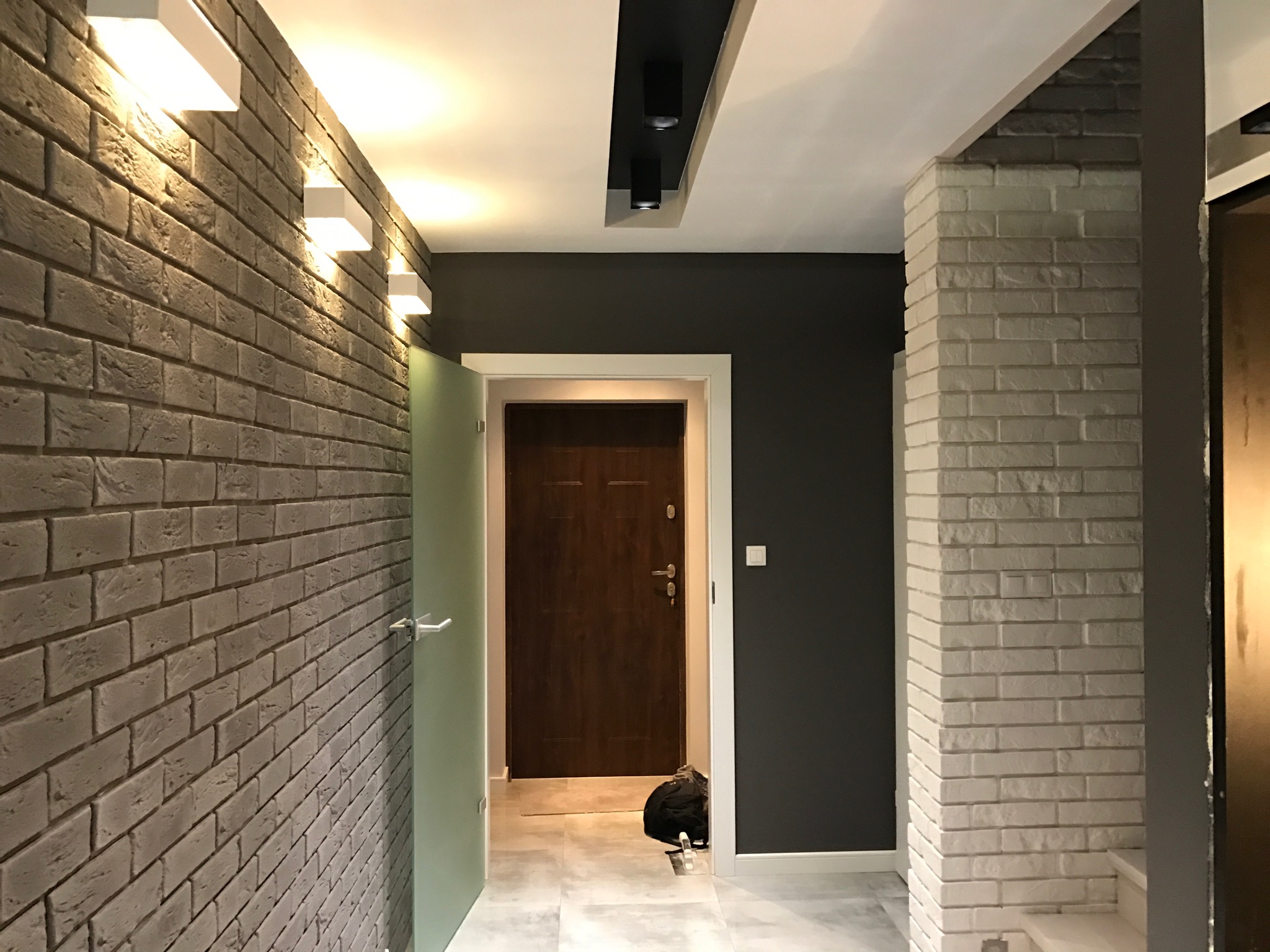 Korytarz z ceglaną ścianą w odcieniach szarości, oświetlony białymi lampami ściennymi, ciemnoszara ściana z białymi listwami przypodłogowymi, drewniane drzwi wejściowe i fragment kolumny z białej...